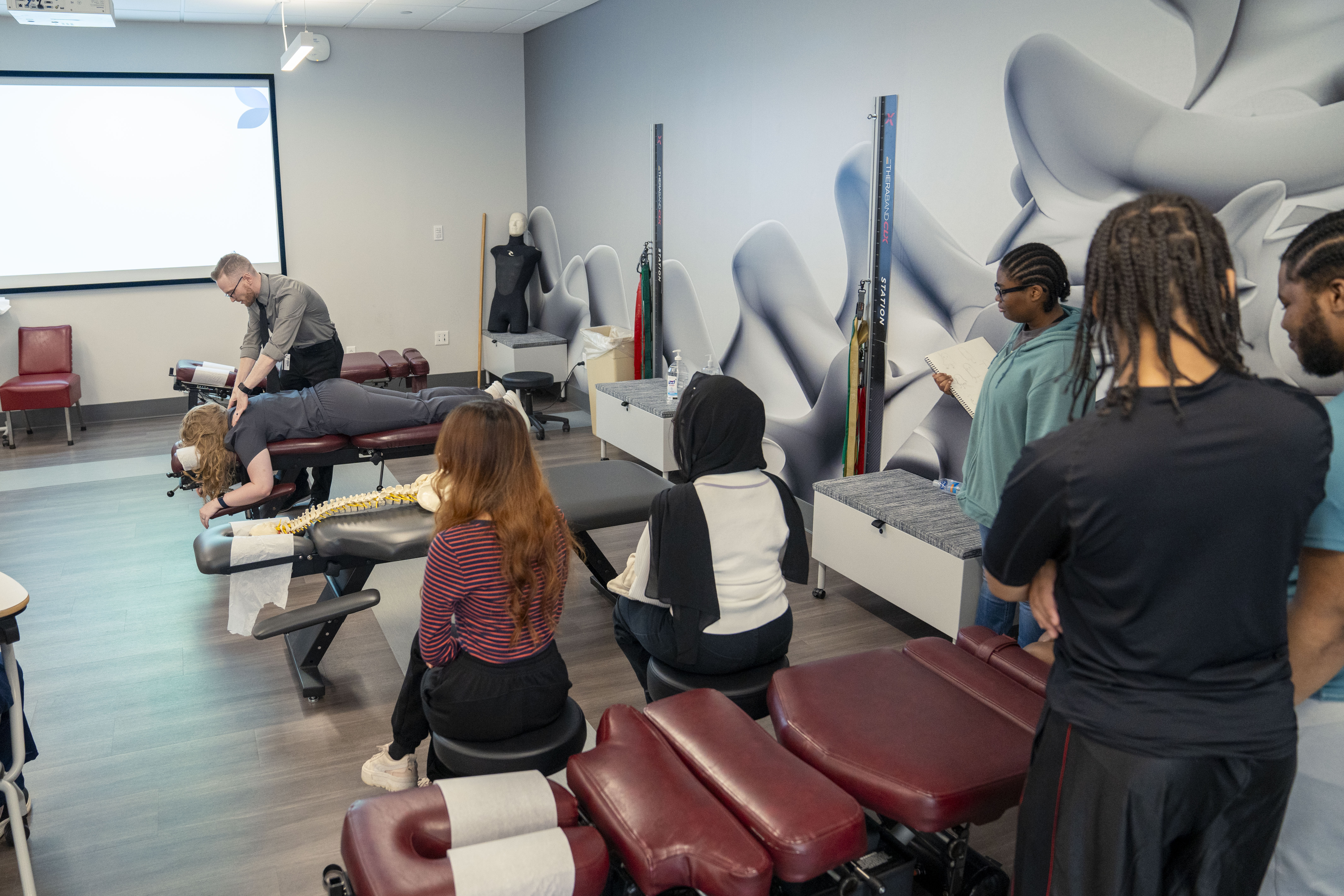 students gather around chiropractic tables watching a chiropractic instructor work on a DYouville student demonstration patient