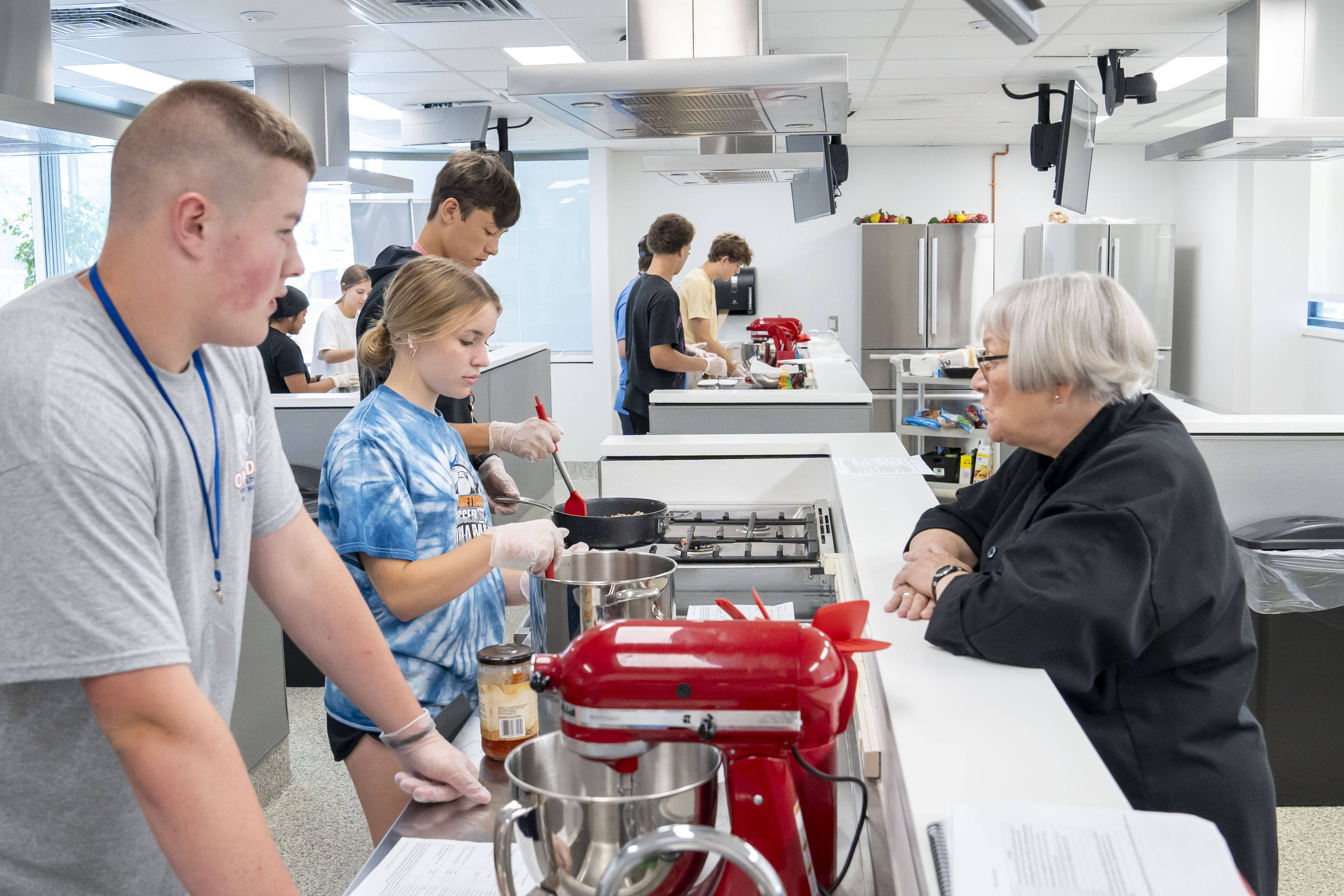 high school students stand inside a industrial kitchen. one is stirring a pot on a stove and another is cooking ground meat in a pan.