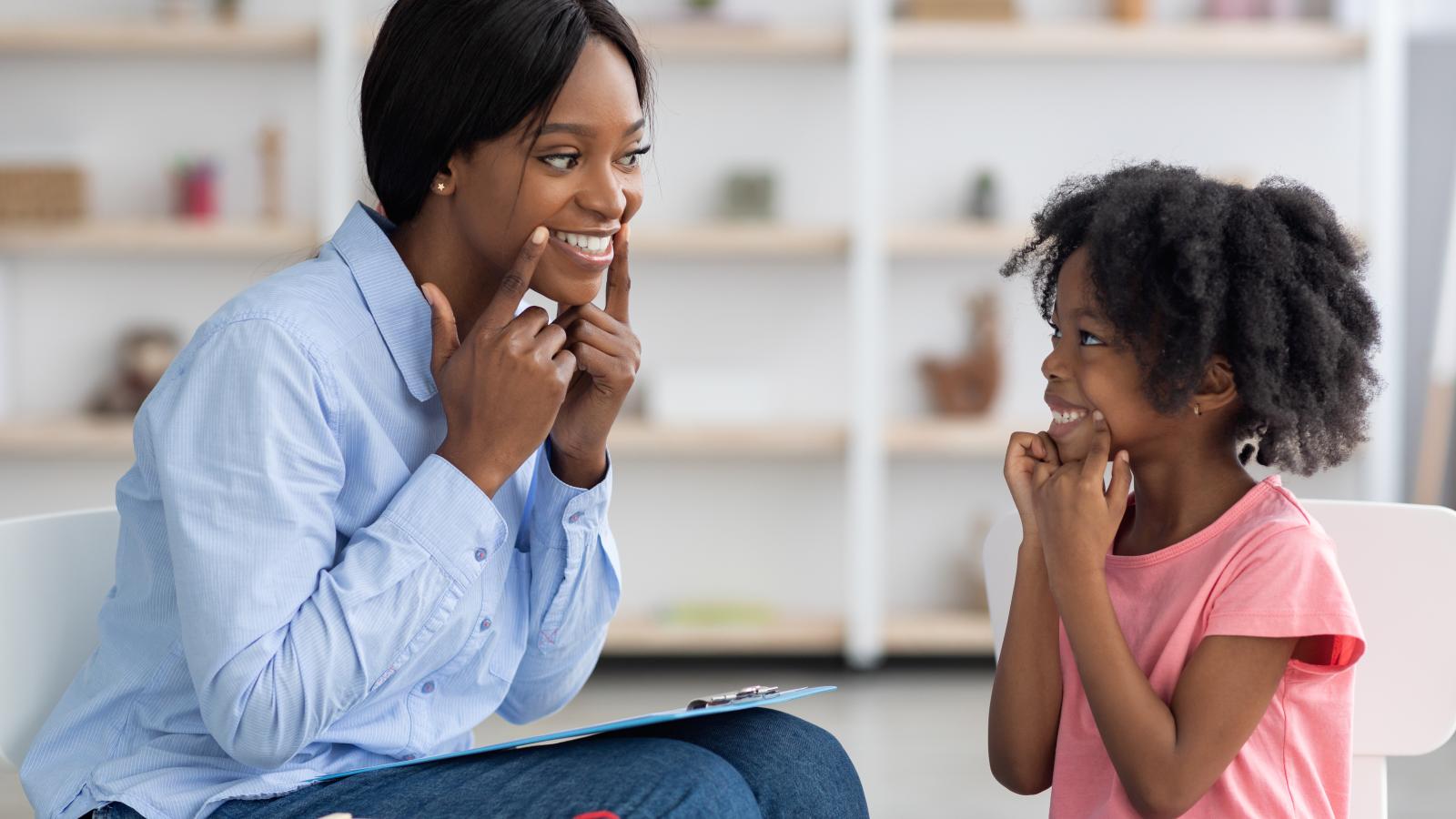 young woman smiling at a child. Both are pointing to the corners of their mouths.