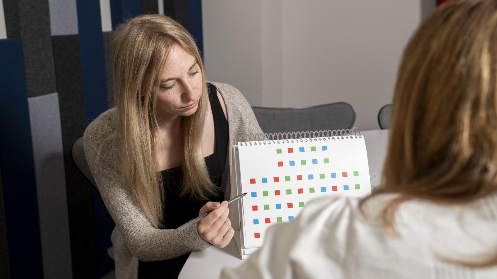 Woman points to a paper with many colored squares. You are looking over the shoulder of another woman.