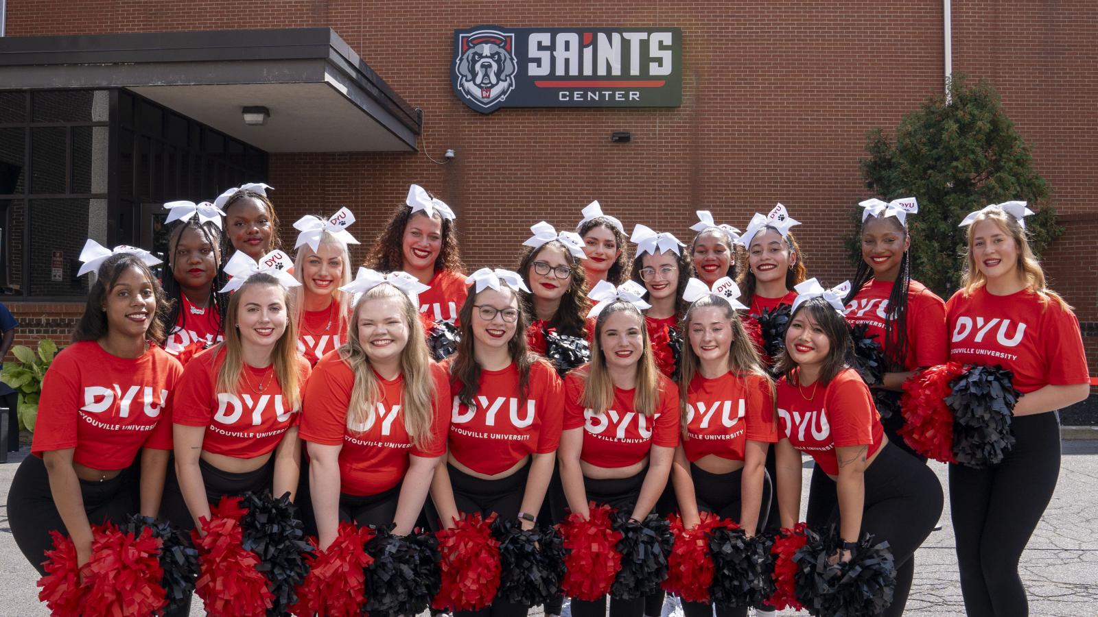 A group of cheerleaders with DYU shirts stand together for a group photo in front of a building displaying a sign that reads: Saints Center