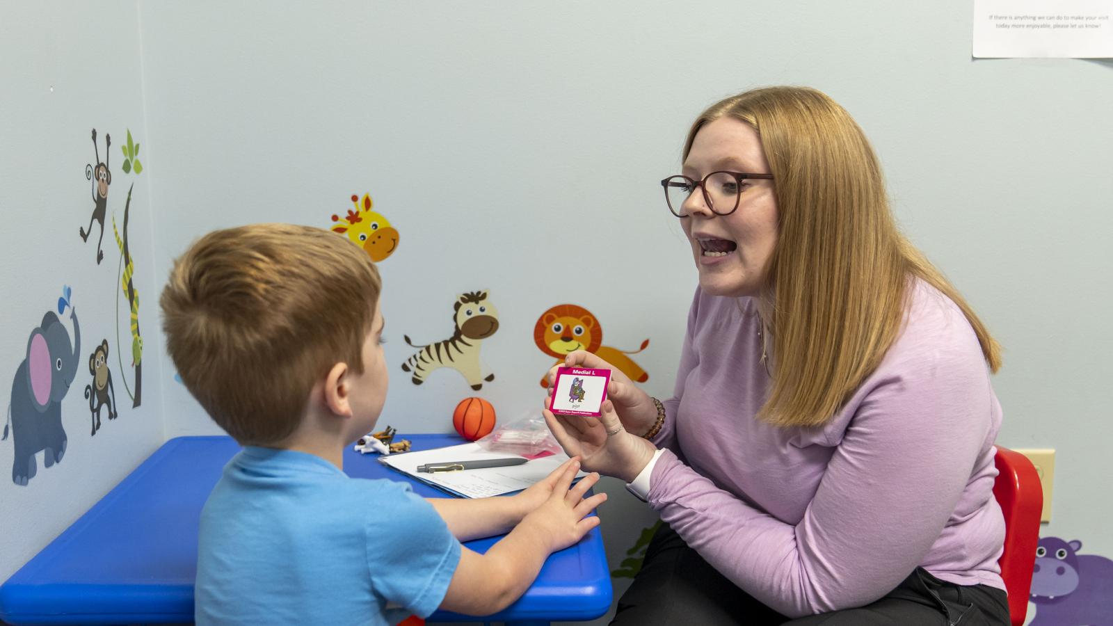 Woman sits at a child size table with young boy. She holds a card with a photo and is shaping her mouth to make an "L" sound.