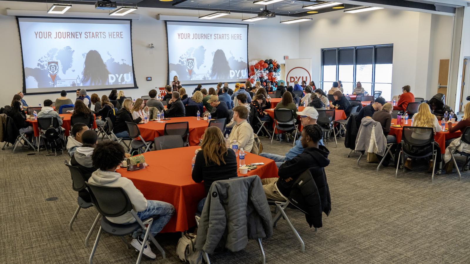 large room filled with round tables. people sit around the tables looking at presentation at the front of the room