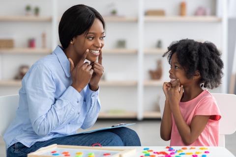 young woman smiling at a child. Both are pointing to the corners of their mouths.