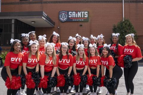 A group of cheerleaders with DYU shirts stand together for a group photo in front of a building displaying a sign that reads: Saints Center