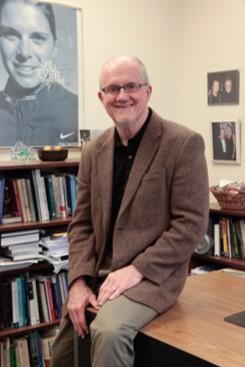 Dr. Donald Sabo, man in brown coat and jacket sits on a desk smiling at the camera