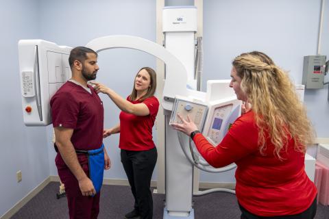 Three people in a room with x-ray machine. Man stands for an x-ray. one woman is operating the machine while the other is pointing and teaching.