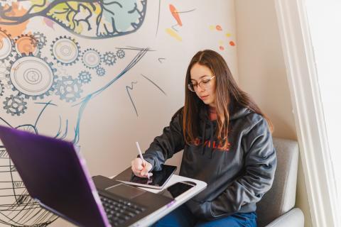 woman writing on tablet with laptop sitting on table in front of her