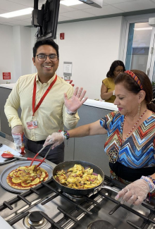 man smiles and waves at the camera. Woman next to him stands in front of a stove cooking inside the D'Lish kitchen