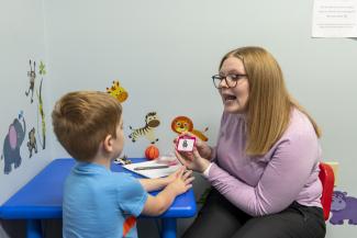 Woman sits at a child size table with young boy. She holds a card with a photo and is shaping her mouth to make an "L" sound.