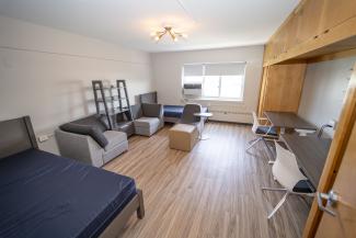 Double room on fourth floor of Madonna Hall. You see two bedframes, a bookshelf, three chairs, two desks and chairs, and a built in cabinet.