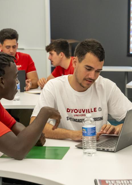 Male students in a classroom discussing classwork and referencing a laptop