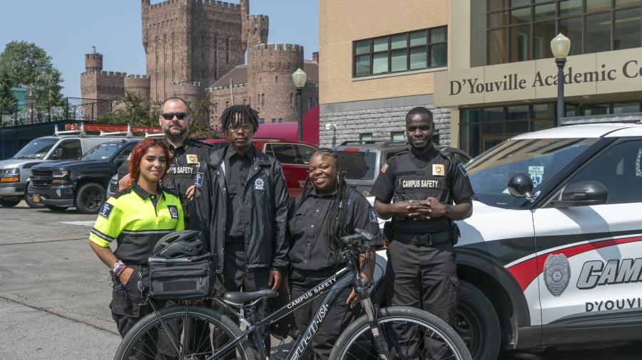 A group of five campus safety officers stands in front of a campus safety patrol car. In the background you can see the D'Youville Academic Center and the Connecticut Street Armory. 