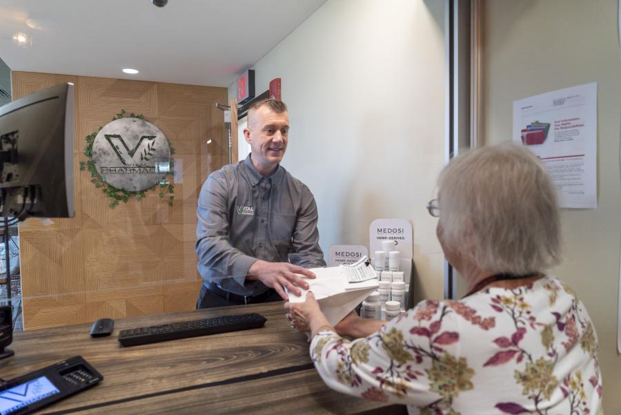 Pharmacist hands a package to a woman over a counter. You see a Vital Pharmacy sign in the background.