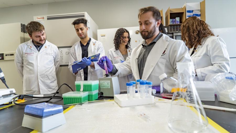 Four students in white coats in a lab space. One student is holding a vial over a vortex mixer. vial of fluid over 