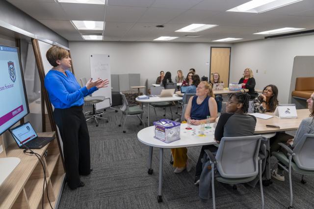 woman stands at the front of a classroom speaking to a large group.