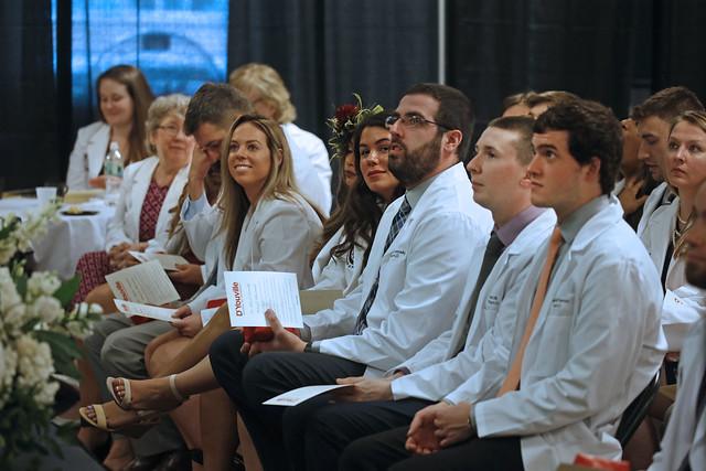 About a dozen young men and women wearing white coats and dresses or ties, seated in rows and looking at something out of frame