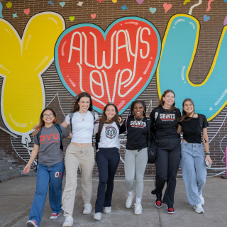 six students walking arm in arm in front of a mural. The mural reads: Always Love You!