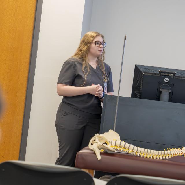 woman in grey scrubs presents next to a lectern in a classroom. In front of her you see a model of the spine.